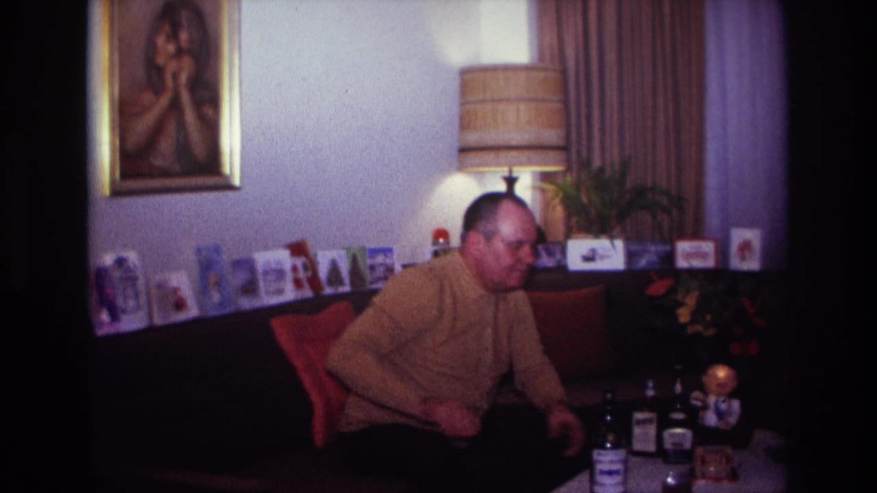 1967 Rockaway Beach home-movie frame of a man seated in a living room with bottles, cards, a lamp, and framed wall art behind him.