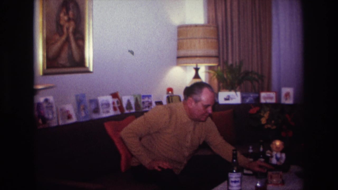 Seated man reaches toward a coffee table with bottles and glassware.