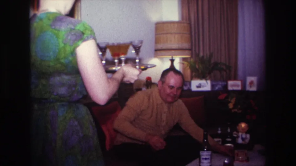 A woman in a green and purple patterned dress serves drinks to a seated man in a 1967 Rockaway Beach living room.