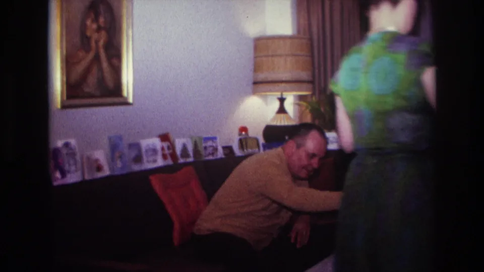 The woman stands near the couch as the seated man reaches toward the table in the Rockaway Beach living room home movie.