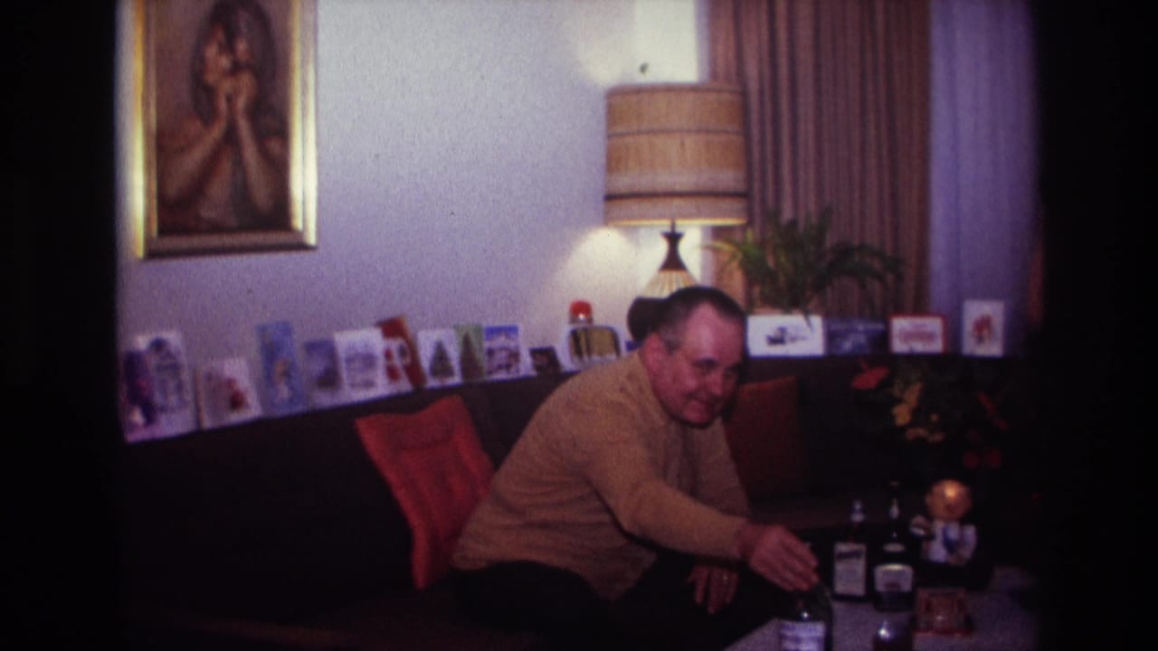 In a 1967 Rockaway Beach home interior, a man sits on a couch near bottles, holiday cards, a lamp, and a framed portrait.