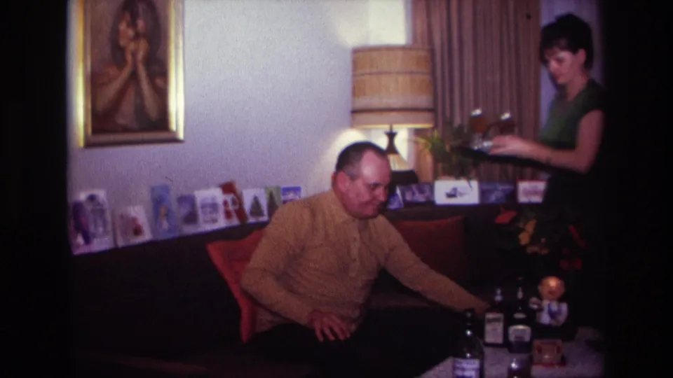 A woman carrying drinks steps into the 1967 living room while the seated man reaches toward the table with bottles.