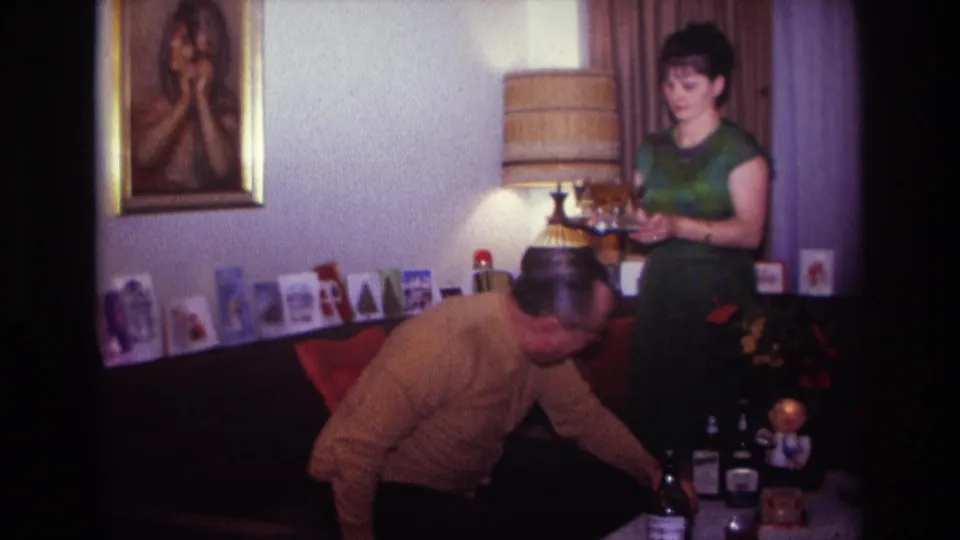 The woman stands holding drinks as the man leans forward near bottles, holiday cards, a lamp, and a couch in the home interior.