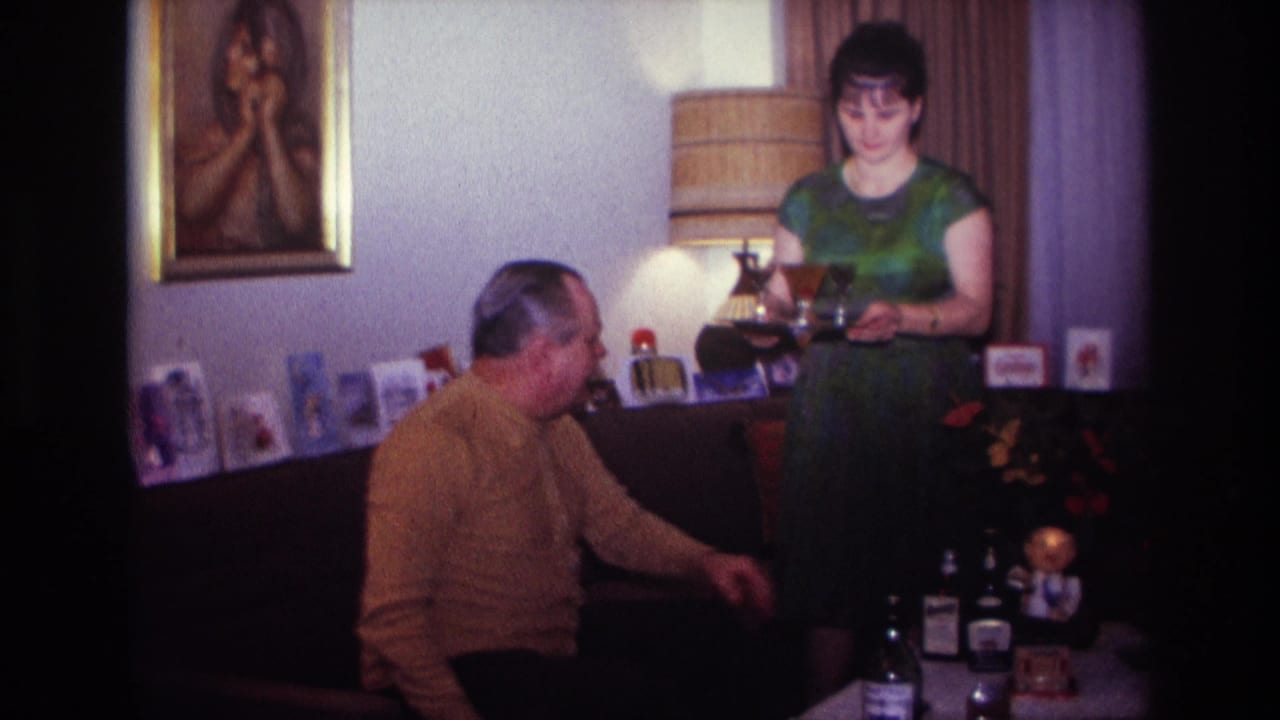 1967 Rockaway Beach home-movie frame showing a woman serving martinis to a seated man in a warmly lit living room.