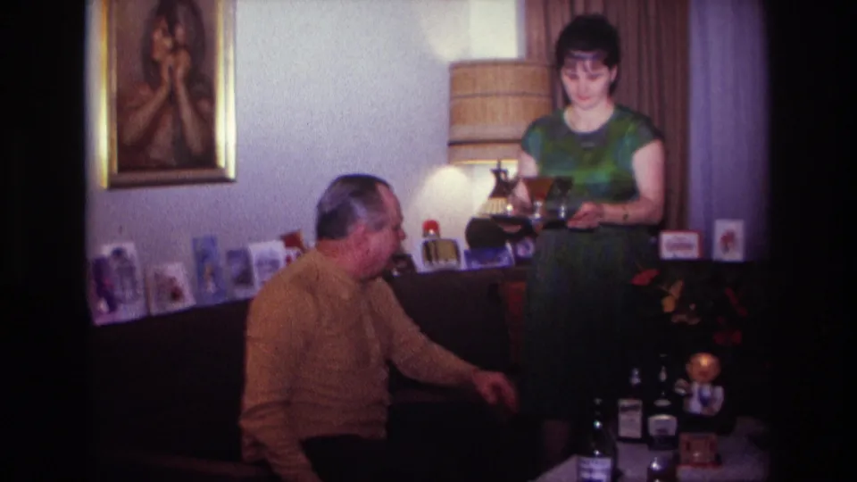 1967 Rockaway Beach home-movie frame showing a woman serving martinis to a seated man in a warmly lit living room.