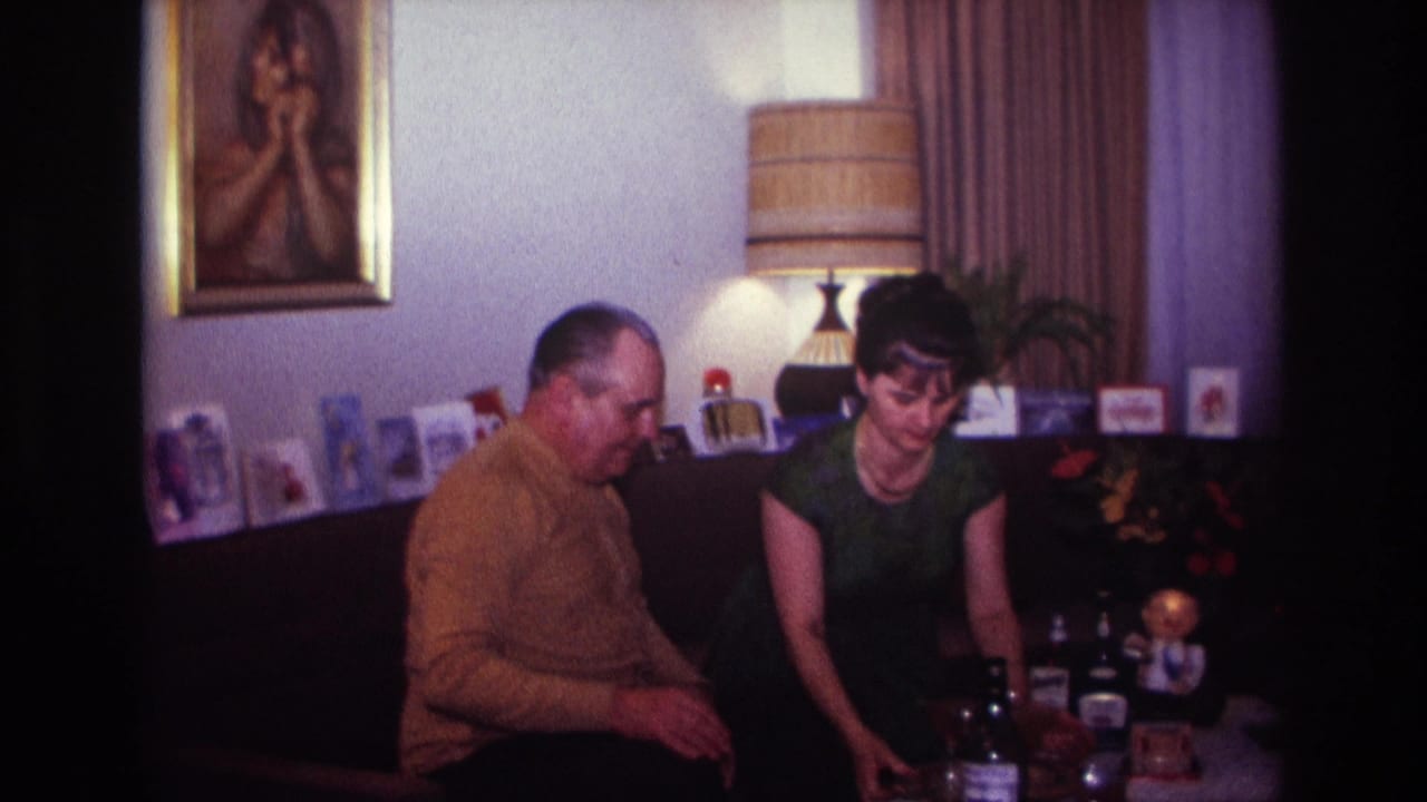 1967 Rockaway Beach home-movie frame of a man in a mustard sweater and a woman in a green dress seated in a living room.