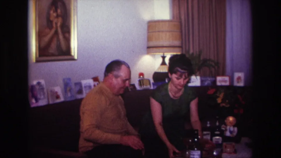 1967 Rockaway Beach home-movie frame of a man in a mustard sweater and a woman in a green dress seated in a living room.
