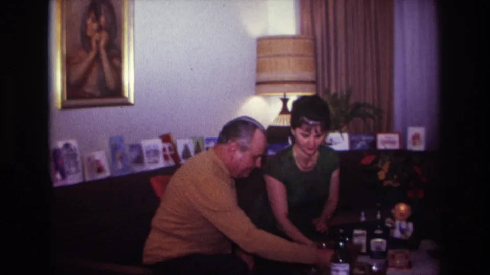 A 1967 home-movie frame shows the couple on a couch as the man leans toward drinks in a mid-century living room.