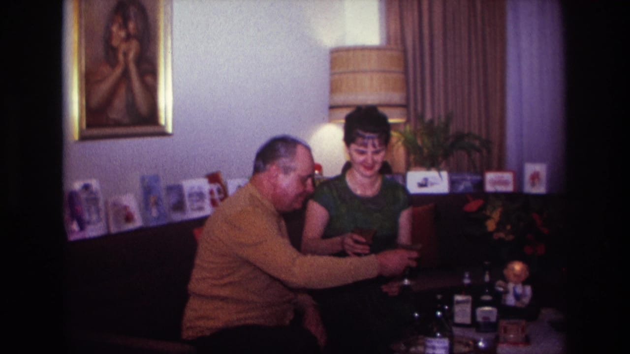 In a 1967 Rockaway Beach home interior, two adults sit close together with drinks as cards, a lamp, and a framed portrait appear behind them.