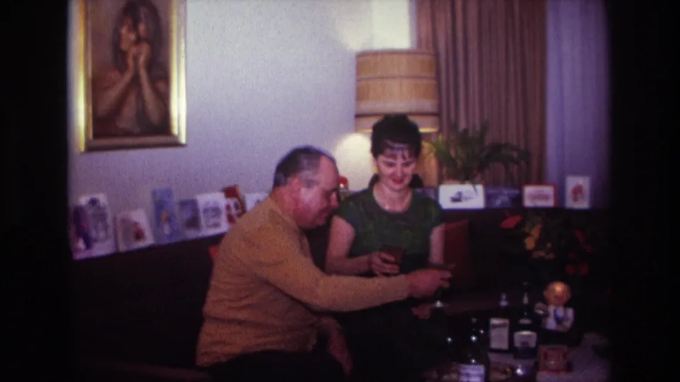 In a 1967 Rockaway Beach home interior, two adults sit close together with drinks as cards, a lamp, and a framed portrait appear behind them.