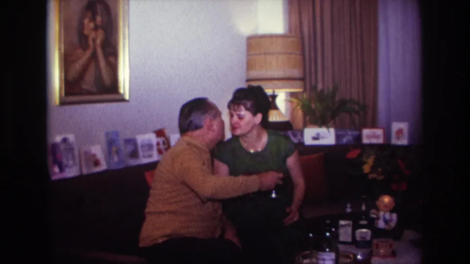 The man leans in close to the woman during an intimate living-room moment in the 1967 Rockaway Beach home-movie clip.