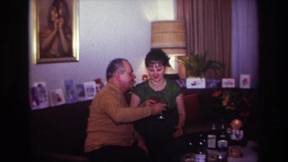 Two adults remain seated close together after a toast in a 1967 Rockaway Beach living room with cards, bottles, flowers, and warm lamp light.