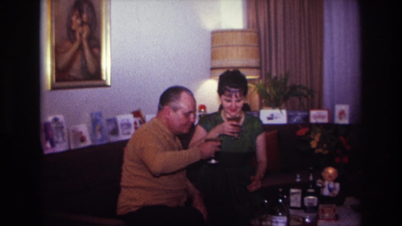 In a 1967 Rockaway Beach home-movie frame, an adult couple sit in a living room and lift wine glasses beside tabletop bottles and decor.