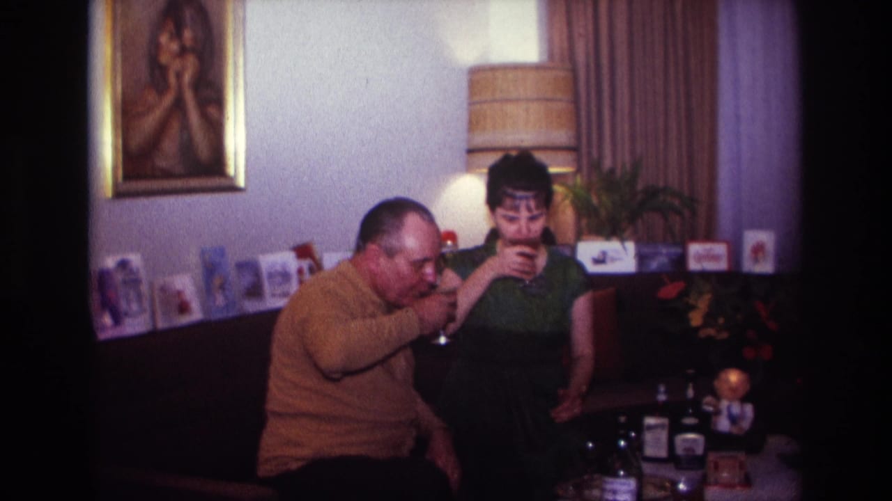 Man and woman sip from wine glasses while seated beside a table with bottles.