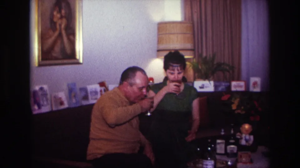 The couple sip wine in a private Rockaway Beach living room, with a lamp, framed portrait, curtains, and bottles visible around them.