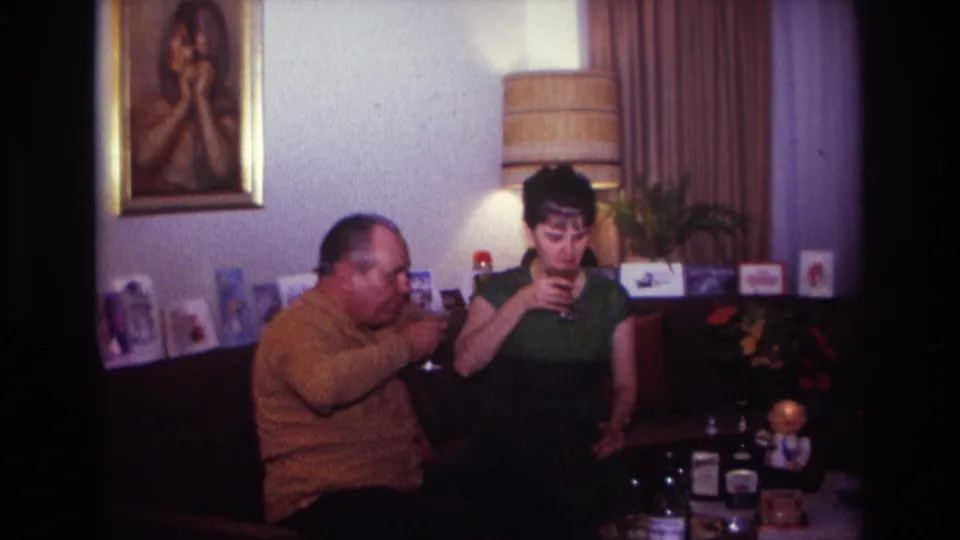 A woman in a green dress holds her glass while a man in a mustard sweater drinks beside her in the 1967 home interior.