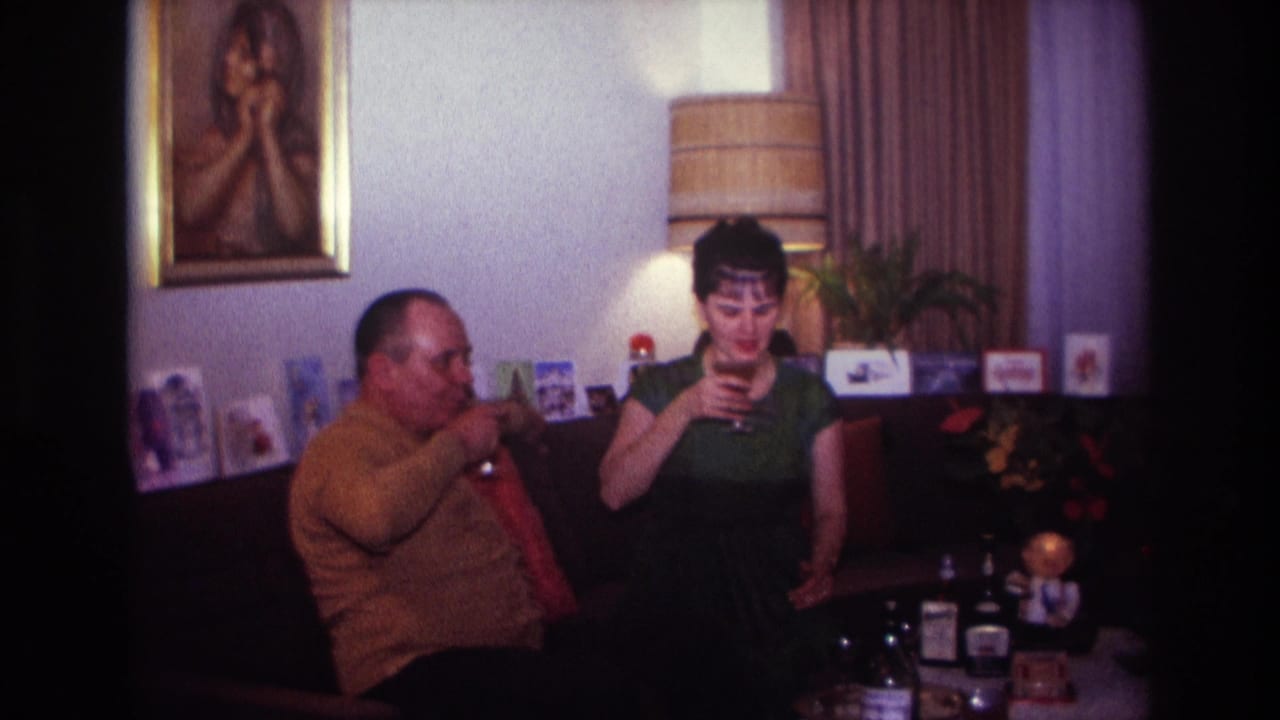 1967 Rockaway Beach home-movie frame of two adults on a couch, with bottles, lamp, curtains, and a framed portrait behind them.