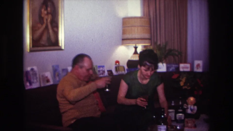 1967 Rockaway Beach living-room frame of a man gesturing toward a woman holding a drink, with bottles, lamp, portrait, and curtains behind them.