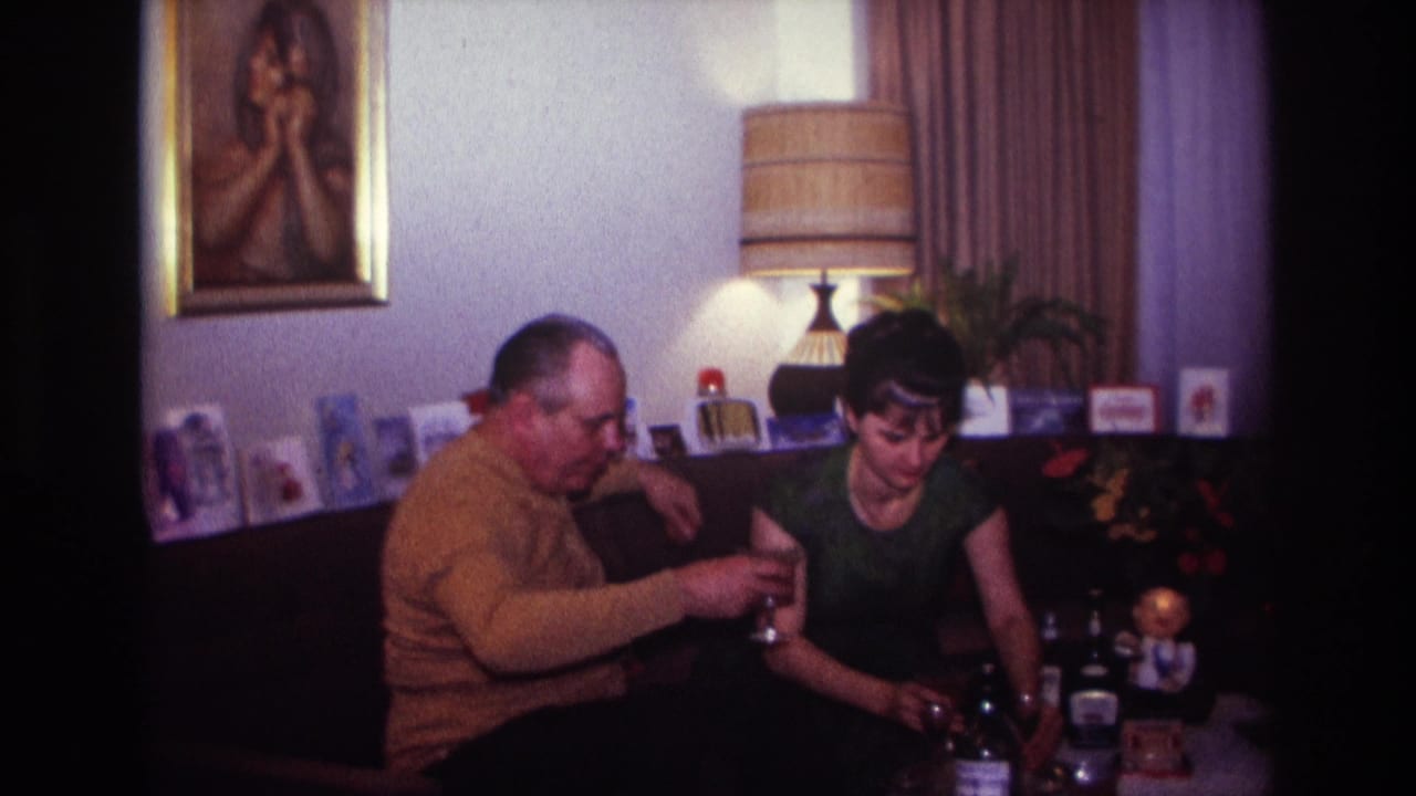 1967 Rockaway Beach home-movie frame of two adults on a couch beside bottles, glasses, greeting cards, a lamp, and a framed portrait.