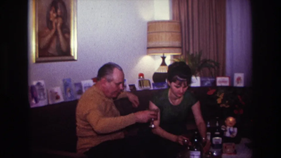 1967 Rockaway Beach home-movie frame of two adults on a couch beside bottles, glasses, greeting cards, a lamp, and a framed portrait.