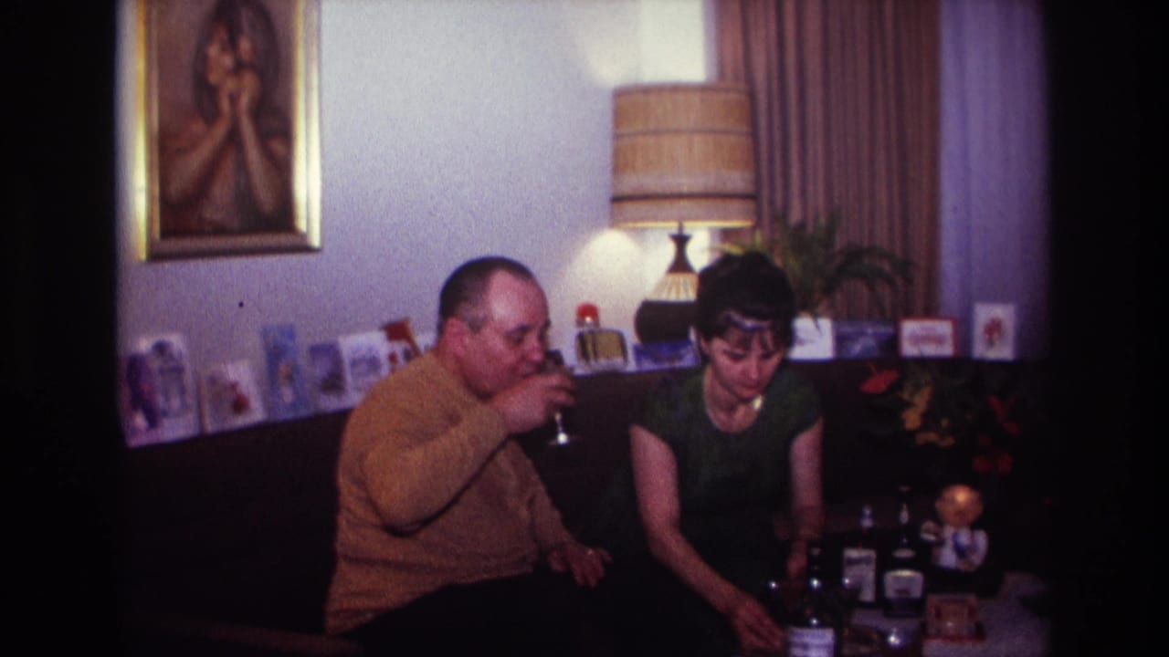 1967 Rockaway Beach interior frame showing a man drinking beside a seated woman, with cards, lamp, portrait, bottles, and glassware visible.