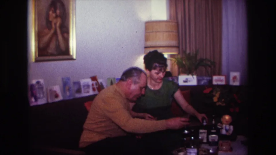 1967 Rockaway Beach living-room frame with a woman smiling and a man leaning toward bottles, glasses, and small items on the table.