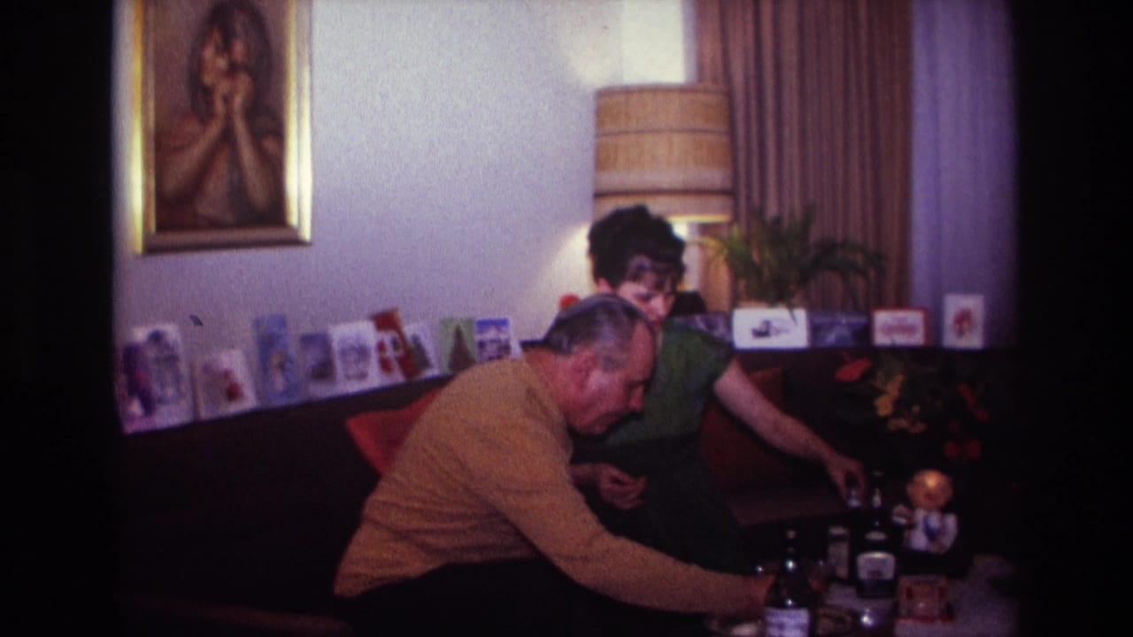 1967 Rockaway Beach home-movie frame showing two adults on a living-room couch, with greeting cards, a lamp, and drink bottles nearby.