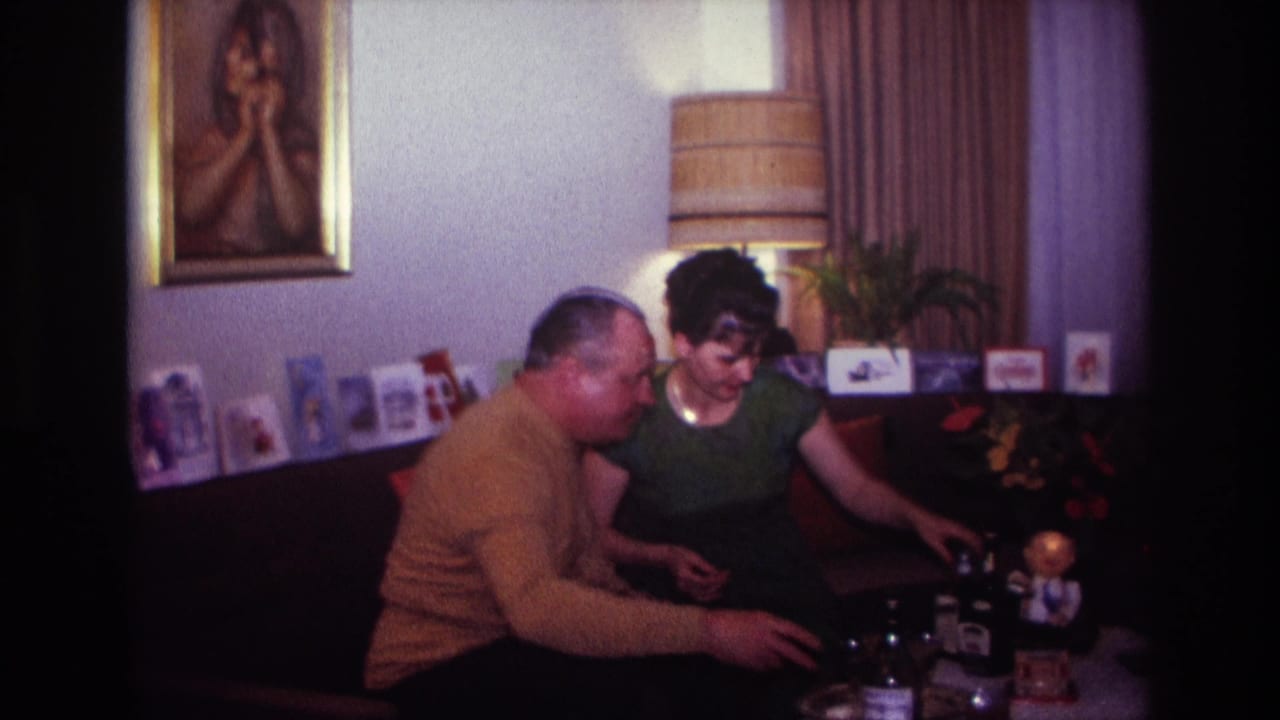 Man and woman sit together as the woman reaches toward a bottle on the table.