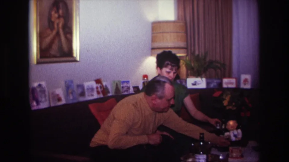 1967 Rockaway Beach interior frame with two adults seated on a couch, surrounded by table drinks, flowers, greeting cards, curtains, and a framed portrait.