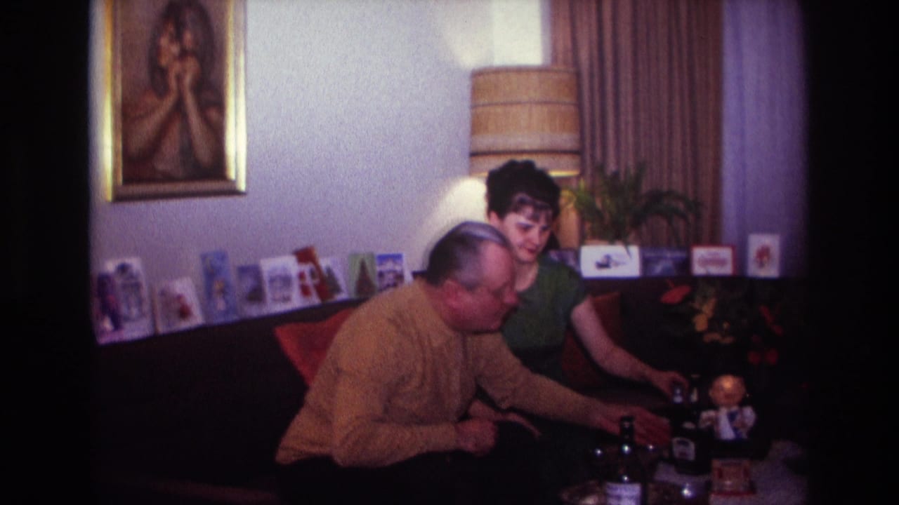 1967 Rockaway Beach home-movie frame of a man in a mustard sweater and a woman in green seated on a couch near table objects.