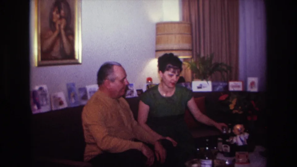 1967 Rockaway Beach living-room frame with two adults seated close together, a lamp, greeting cards, and wall portrait visible.