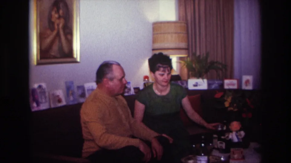 Final 1967 home-movie frame of two adults in a Rockaway Beach living room, with bottles and a small toy on the table.