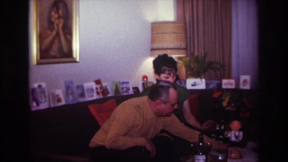 1967 Rockaway Beach home-movie frame with the man leaning forward near bottles and glasses as the woman sits on the sofa.