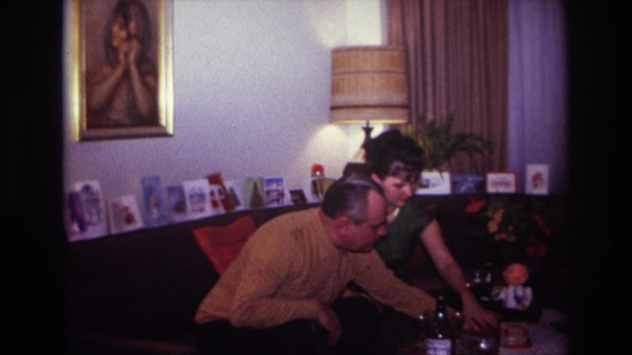 In a 1967 Rockaway Beach home-movie frame, an adult couple sit on a dark sofa near a lamp, portrait and greeting cards.
