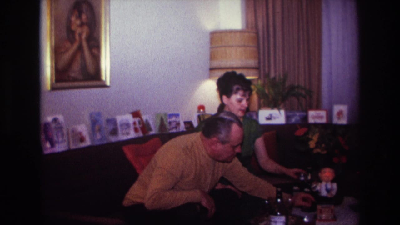 1967 Rockaway Beach home-movie frame of two adults in a living room, leaning toward a coffee table with a small decoration.