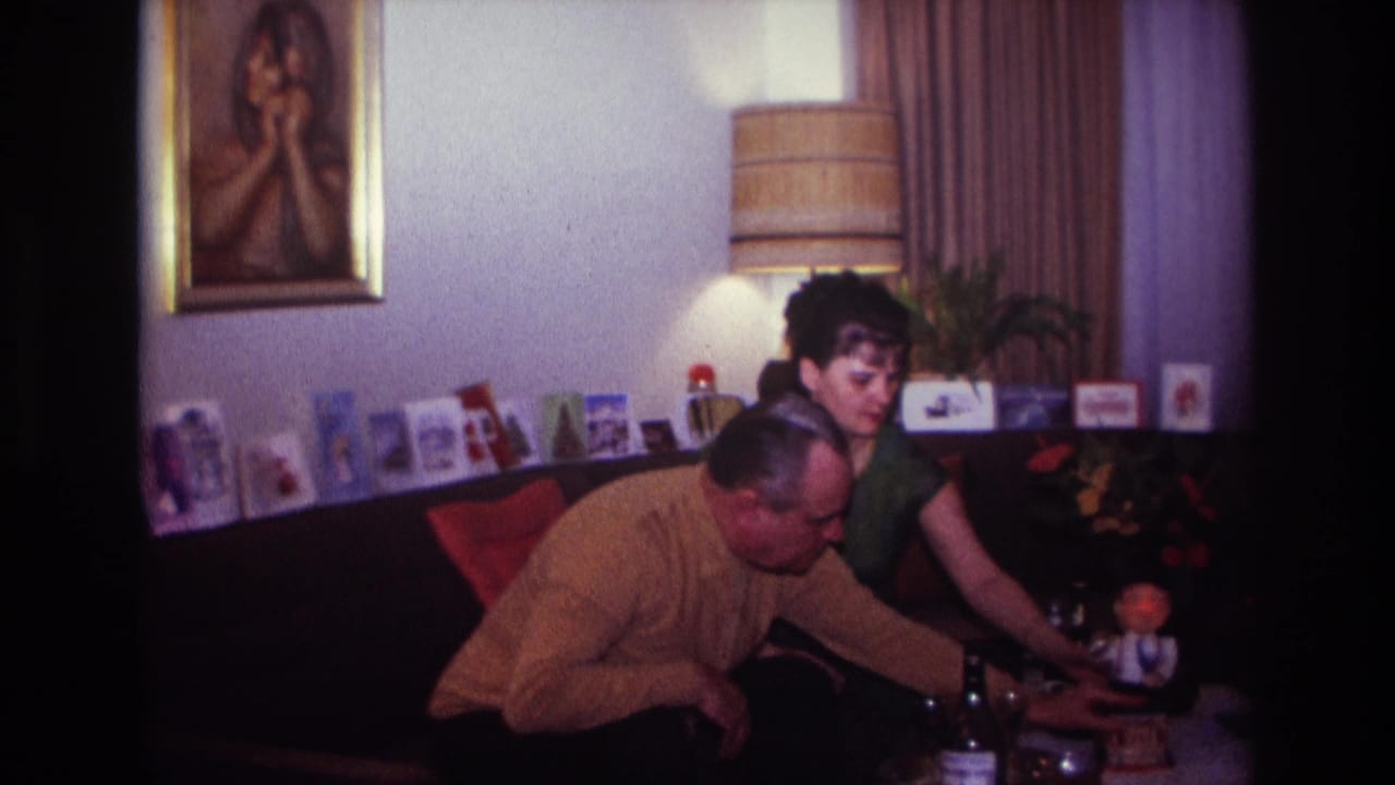 Woman reaches toward a small figurine as a man leans across the coffee table in a card-lined living room.