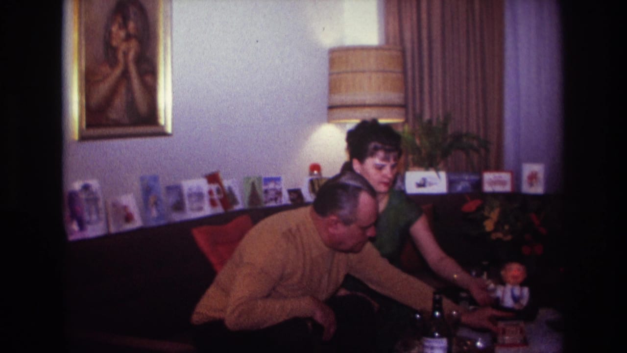 1967 Rockaway Beach home-movie frame of a man in a mustard sweater and woman in green seated beside a doll in a decorated living room.
