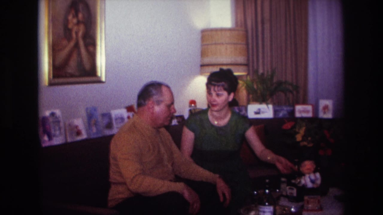 In a 1967 Rockaway Beach home interior, a man in a mustard sweater and a woman in a green dress sit on a couch near tabletop objects.
