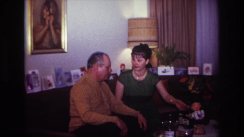 In a 1967 Rockaway Beach home interior, a man in a mustard sweater and a woman in a green dress sit on a couch near tabletop objects.