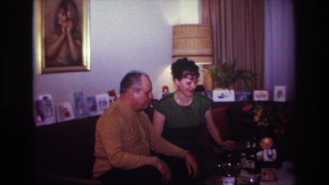 Seated man looks toward a woman beside a table with small objects.