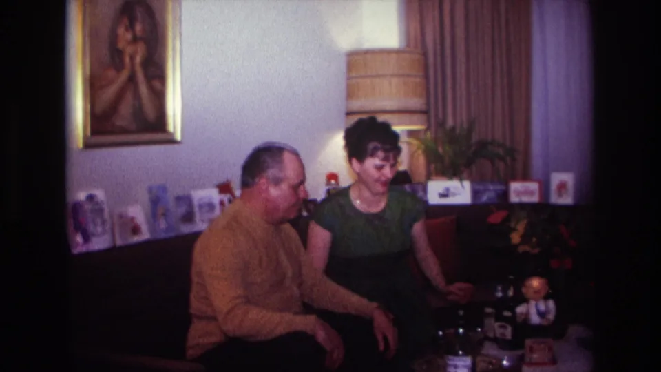 The seated pair continue talking in the 1967 living room, with the woman turned toward the table and a small figurine visible nearby.