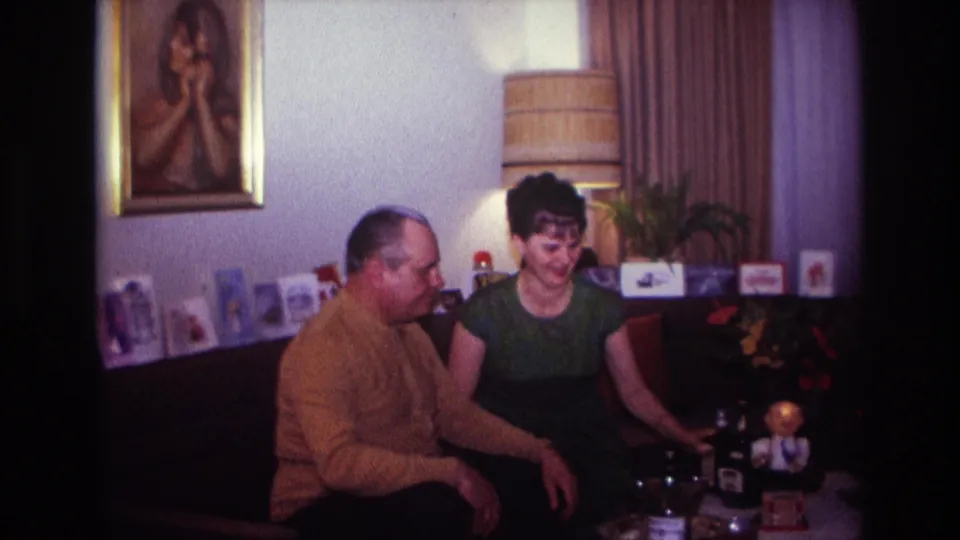 The woman smiles in the 1967 Rockaway Beach living room as the man sits beside her, with framed art, lamp, and wall cards behind them.