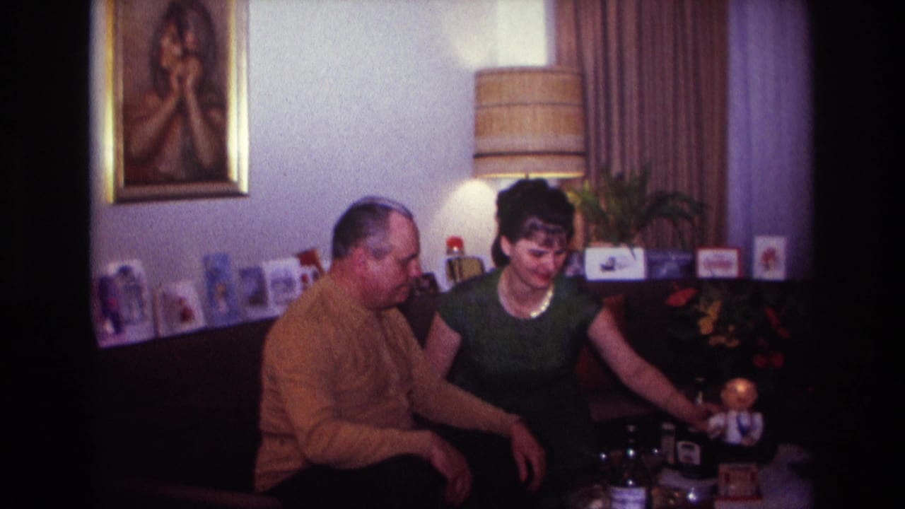1967 Rockaway Beach home-movie frame of a man in a mustard sweater and a woman in green seated beside a table with a toy figure.