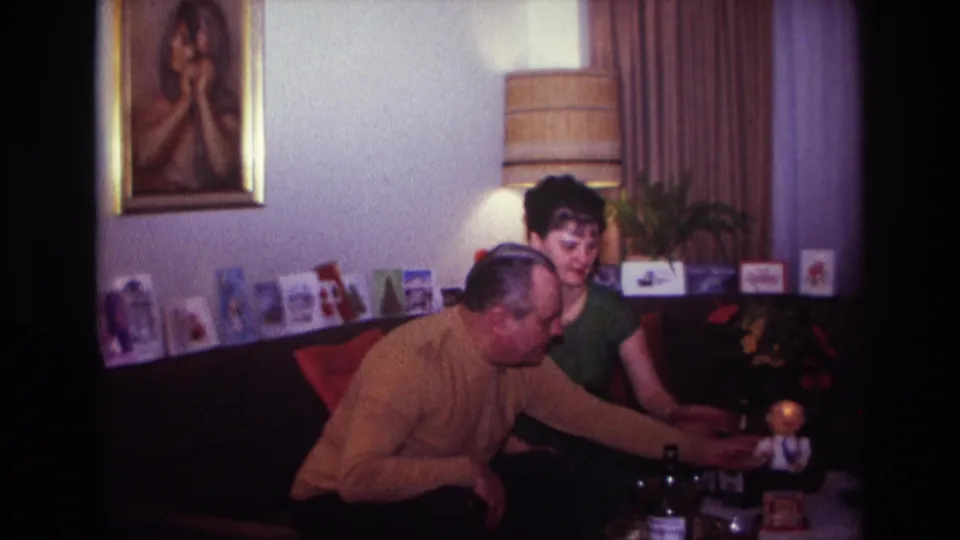1967 Rockaway Beach frame of a man leaning across the living room table toward a small toy figure while a woman sits beside him.