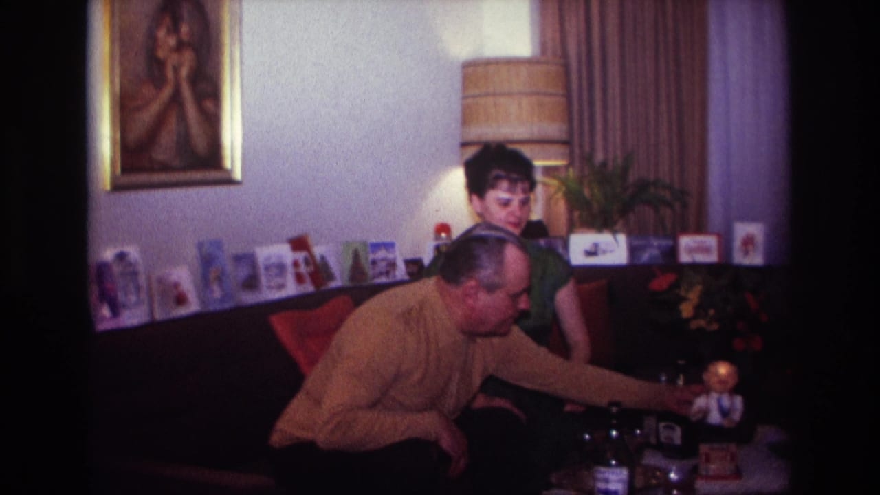 1967 Rockaway Beach home-movie frame of a man in a mustard sweater reaching toward a tabletop toy while a woman in green watches.
