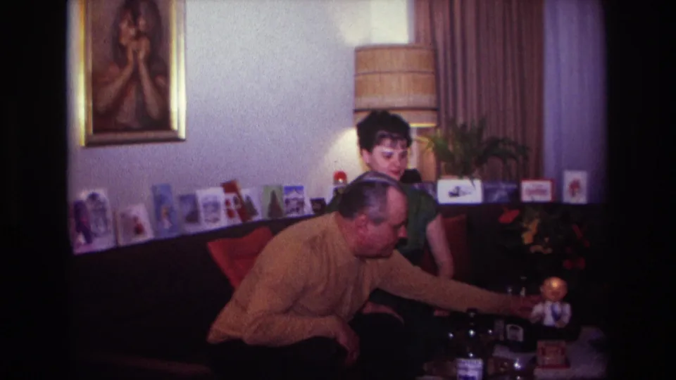 1967 Rockaway Beach home-movie frame of a man in a mustard sweater reaching toward a tabletop toy while a woman in green watches.