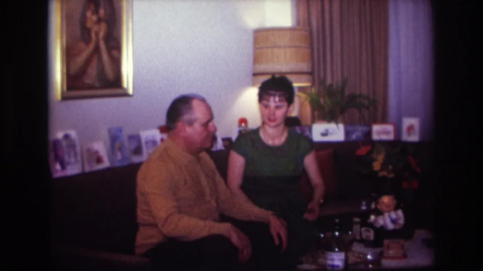 1967 Rockaway Beach frame of two adults seated together on a couch with a coffee table, toy, cards, lamp, and wall art visible.