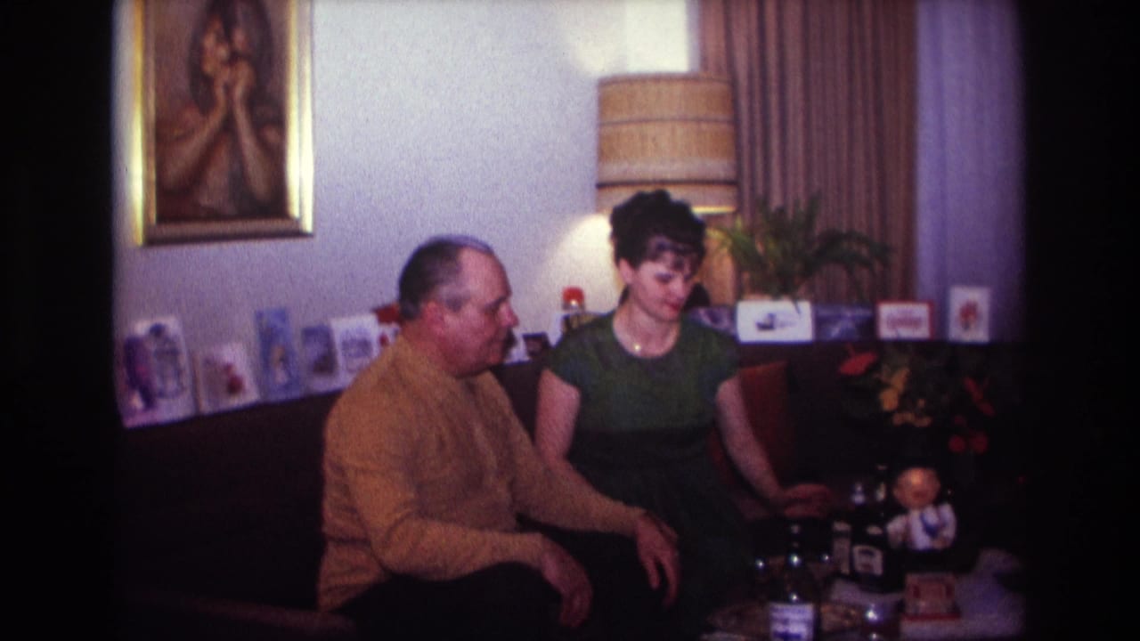 In a 1967 Rockaway Beach home-movie frame, a man in a mustard sweater sits beside a woman in a green dress in a living room.