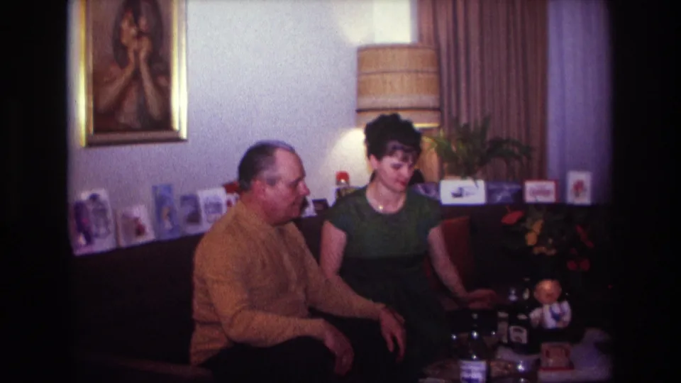 In a 1967 Rockaway Beach home-movie frame, a man in a mustard sweater sits beside a woman in a green dress in a living room.