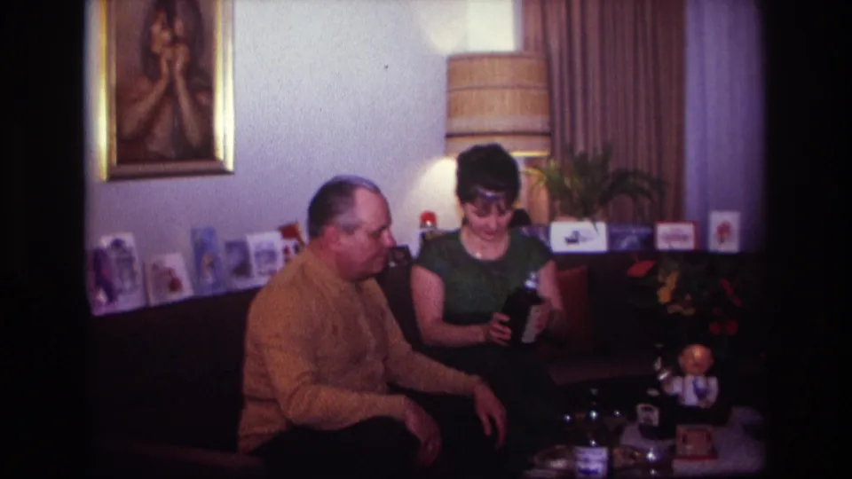 The woman in a green dress holds a dark bottle as the man beside her watches in the Rockaway Beach living room scene.
